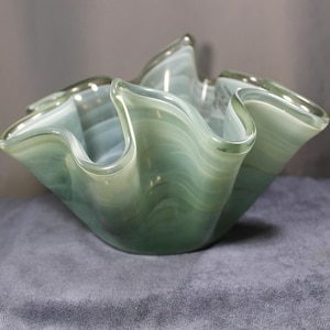 May include: A decorative glass bowl with a ruffled, wave-like rim. The bowl is a pale green color with white swirls throughout the glass. The bowl has a wide, open shape.