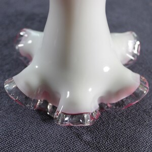 May include: A white glass vase base featuring a flared, ruffled edge with a translucent pink trim. The smooth, opaque white body transitions into a decorative, crimped bottom rim that displays a clear and pink glass finish at the very edge.