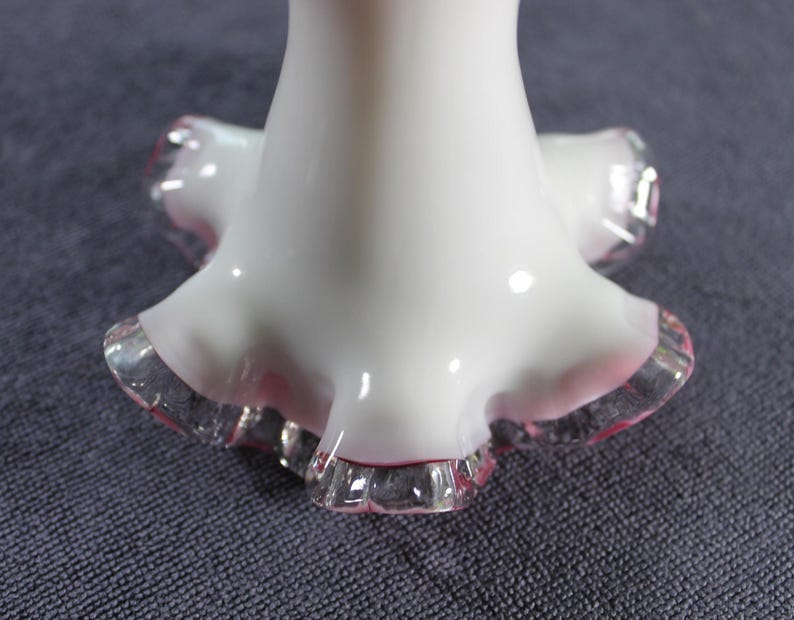 May include: A white glass vase base featuring a flared, ruffled edge with a translucent pink trim. The smooth, opaque white body transitions into a decorative, crimped bottom rim that displays a clear and pink glass finish at the very edge.