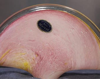 May include: A Murano glass bowl with a pink and yellow color scheme. The bowl has a rounded shape with a clear rim. A small, oval sticker with the text "Lavorazione Arte Murano Made in Italy" is affixed to the bowl.