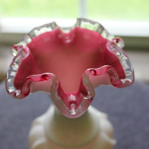 May include: A glass vase featuring a wide, ruffled opening with a vibrant pink interior that transitions into a lighter, opaque white base. The rim of the vase is finished with a clear, smooth glass edge, highlighting the undulating, wavy shape.