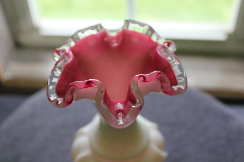 May include: A glass vase featuring a wide, ruffled opening with a vibrant pink interior that transitions into a lighter, opaque white base. The rim of the vase is finished with a clear, smooth glass edge, highlighting the undulating, wavy shape.