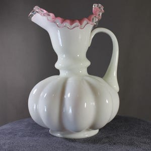 May include: White glass pitcher with a bulbous body and a ruffled pink rim. The pitcher has a curved handle and a spout for pouring. The body of the pitcher has vertical ridges. A decorative piece for serving beverages.