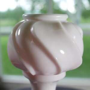 May include: A pale pink milk glass vase featuring a rounded, bulbous body with a distinct swirling, ribbed pattern. The glass has a smooth, glossy finish and a narrow, circular opening at the top, tapering down into a slender, solid base.