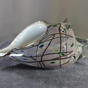 May include: A decorative glass seashell sculpture. The shell is primarily gray with brown, blue, and green accents. The interior is white. The sculpture has a textured surface and is resting on a gray surface.