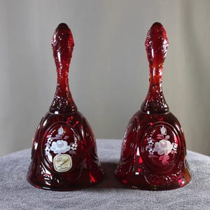 May include: Two ruby red glass bells with long, textured handles. Each bell features a floral design with white and pink accents. The bells are displayed on a gray surface, showcasing their intricate details and rich color.