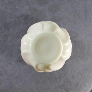 May include: A top-down view of a small, off-white ceramic vessel featuring a smooth, circular central opening surrounded by a series of curved, petal-like ridges that radiate outward, creating a symmetrical, flower-shaped silhouette.