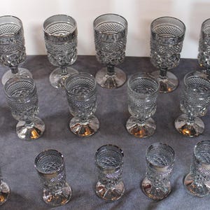 May include: A collection of clear, stemmed glasses with a silver-colored base. The glasses have a textured, diamond-patterned design on the bowl and stem. The glasses are arranged on a gray surface, showcasing their elegant design.