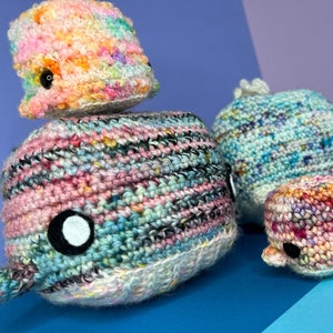 May include: A collection of crocheted whale plushies in various colors, including pink, blue, and green. The whales are all different sizes and have black eyes.