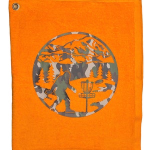 May include: Orange golf towel with a circular design featuring a Bigfoot figure throwing a disc golf disc at a basket in a camouflage pattern.