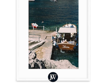 May include: A wooden boat with a white canopy docked at a rocky shore. The boat is labeled "Gloria" and has several people on board. The water is a deep blue and there are other boats in the distance.