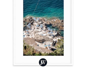 May include: An aerial view of a rocky beach with white lounge chairs and blue and white striped umbrellas. The water is a vibrant turquoise blue.