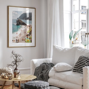 May include: A framed photograph of a beach scene with people swimming and sunbathing. The beach is surrounded by rocky cliffs and the water is a clear turquoise blue. The photo is hanging on a white wall in a living room setting.