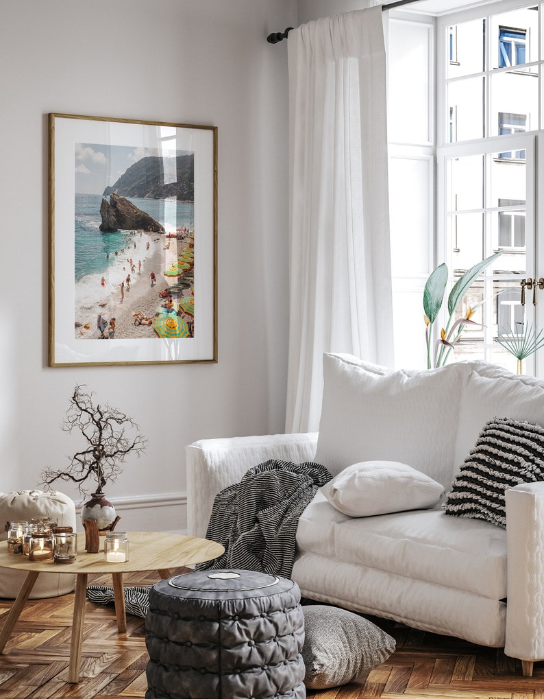 May include: A framed photograph of a beach scene with people swimming and sunbathing. The beach is surrounded by rocky cliffs and the water is a clear turquoise blue. The photo is hanging on a white wall in a living room setting.