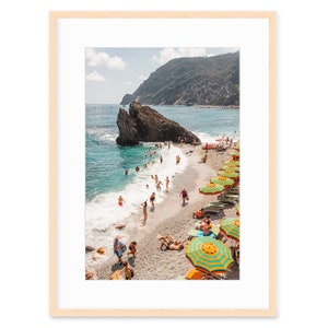 May include: A beach scene with a rocky cliff, turquoise water, and a sandy shore. People are swimming and sunbathing under colourful striped umbrellas.