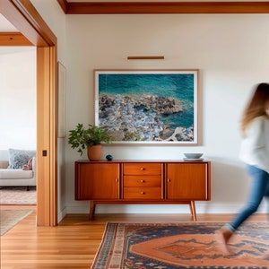 May include: A mid-century modern style wooden cabinet with three drawers and two doors. The cabinet is in a hallway with a rug on the floor and a framed photograph of a seascape on the wall.