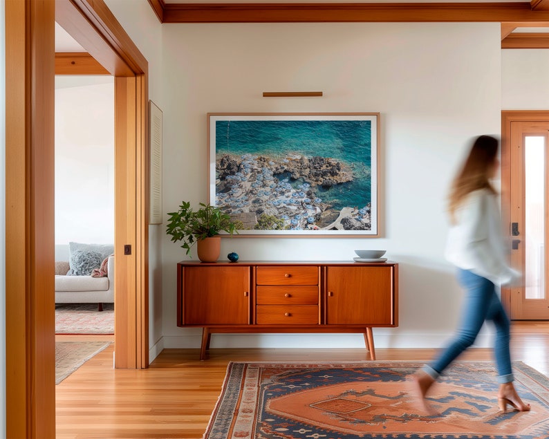 May include: A mid-century modern style wooden cabinet with three drawers and two doors. The cabinet is in a hallway with a rug on the floor and a framed photograph of a seascape on the wall.