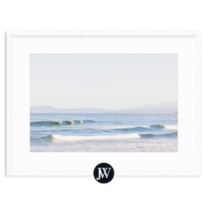 May include: A photograph of a seascape with blue water and white waves crashing on the shore. The horizon is in the distance with a line of mountains in the background. There are several surfers in the water.