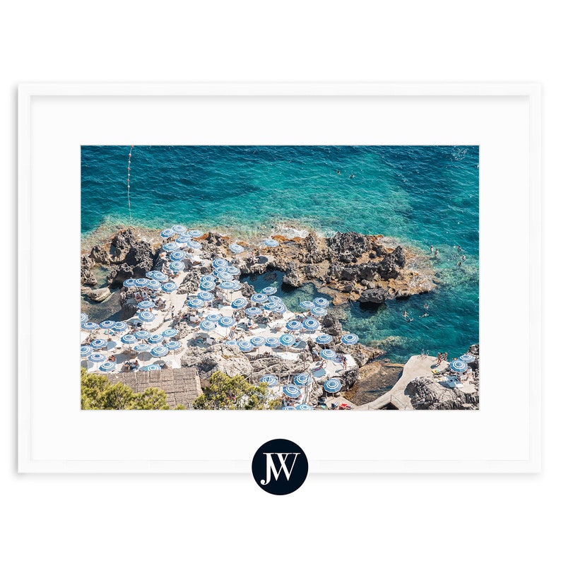 May include: An aerial view of a rocky coastline with a turquoise blue ocean. White and blue striped umbrellas line the shore, creating a pattern on the sand.