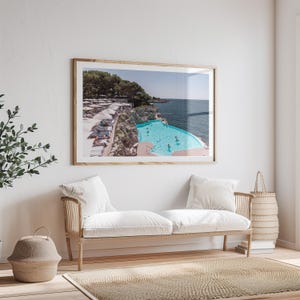 May include: A framed photograph of a swimming pool overlooking the ocean. The pool is surrounded by a rocky cliff and there are white umbrellas and lounge chairs on the cliff. The water in the pool is a bright turquoise blue.