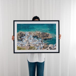 May include: A framed photograph of a beach scene with turquoise water and white umbrellas. The photo is held by a person in front of a white wall.
