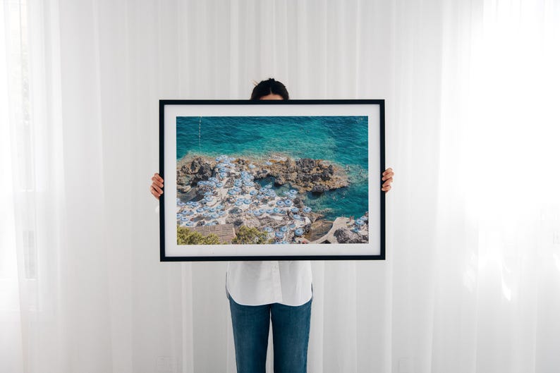 May include: A framed photograph of a beach scene with turquoise water and white umbrellas. The photo is held by a person in front of a white wall.
