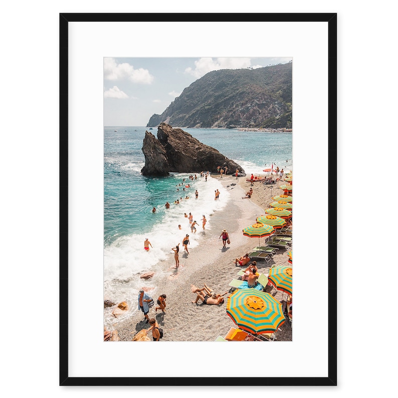 May include: A beach scene with colourful striped umbrellas and people swimming and sunbathing on a sandy beach with a rocky cliff in the background.