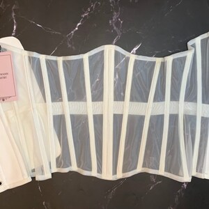 May include: White mesh corset with white boning and metal eyelets. The corset has a label that reads "Shadowrain Corsetory".