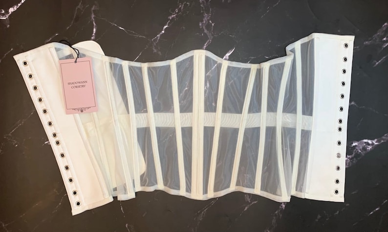 May include: White mesh corset with white boning and metal eyelets. The corset has a label that reads "Shadowrain Corsetory".