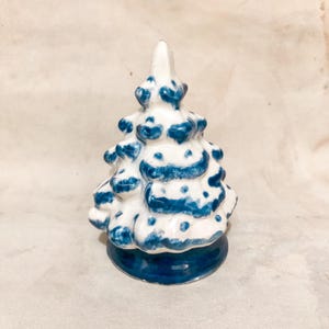 Może przedstawiać: Ceramiczna figurka choinki. Drzewko jest białe z niebieskimi akcentami i niebieską podstawą. Drzewko ma spiczasty wierzchołek i teksturowaną powierzchnię. Figurka jest przedmiotem dekoracyjnym.