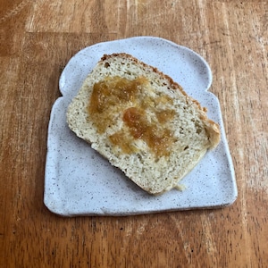 Peut inclure: Une tranche de pain blanc avec une cuillerée de marmelade d'orange sur le dessus, posée sur une assiette en céramique blanche en forme de tranche de pain.
