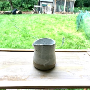 Könnte beinhalten: Ein kleines Milchkännchen aus Keramik mit einer gesprenkelten, cremeweißen Glasur im oberen Bereich und einem naturfarbenen, hellbraunen Tonboden. Das Kännchen hat einen bauchigen Körper, eine weite Öffnung und einen kleinen Ausguss.