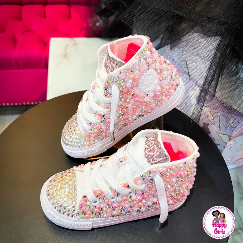 May include: White high-top sneakers with pink and clear rhinestones. The sneakers have a pink and white interior and the words "Happy Girls" are on the side.