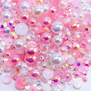 May include: A variety of iridescent pink and white pearl and crystal embellishments. The embellishments are scattered on a white surface.