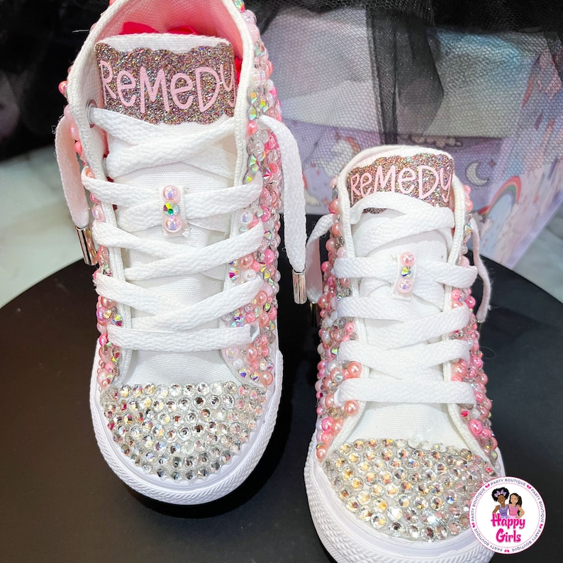May include: A pair of white high-top sneakers with pink and clear rhinestones. The sneakers have the name "Remedy" written on them in pink glitter.