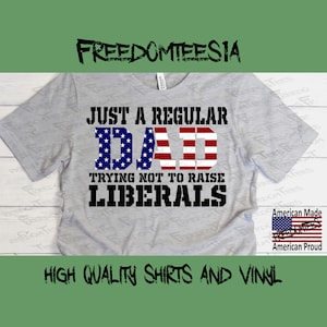 May include: A gray t-shirt with the text "Just a Regular Dad Trying Not to Raise Liberals" in black. The word "Dad" is in red, white, and blue with an American flag design. The text "Freedomteesia" is at the top of the image. The text "American Made" and "American Proud" are at the bottom of the image.