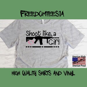 May include: A gray t-shirt with the text "Shoot like a Girl" and a silhouette of a rifle with a pink heart. The text "FREEDOMTEESIA" is at the top of the image. The text "American Made" and "American Proud" are at the bottom of the image.