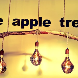 May include: Three light bulbs hang from a rustic branch, suspended by rope. The branch is attached to the ceiling and the bulbs are lit. The text "the apple tree" is written on the wall behind the branch.