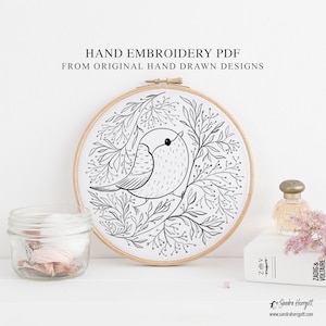 May include: A hand embroidery PDF featuring an original hand-drawn design of a bird surrounded by leaves, displayed in a wooden hoop. The text "HAND EMBROIDERY PDF FROM ORIGINAL HAND DRAWN DESIGNS" is visible. A jar of thread and a perfume bottle are also present.