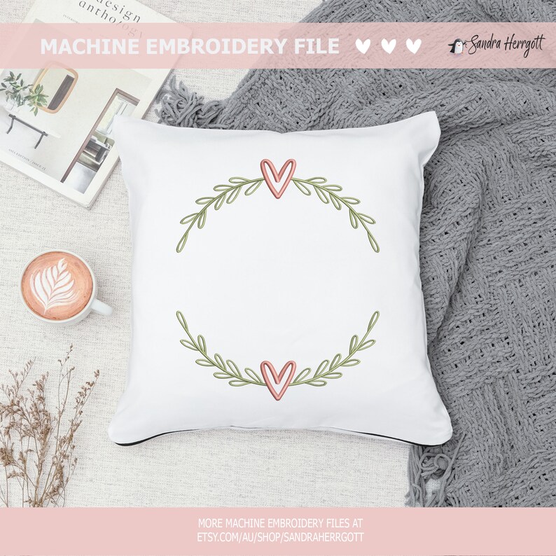 May include: White pillow with a green wreath embroidered with two pink hearts. The wreath is a simple design with leaves and the hearts are filled with a light pink color.