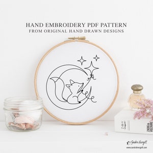 May include: A hand embroidery PDF pattern featuring a minimalist design of a fox sitting on a crescent moon with stars. The design is in black on white fabric, set in a wooden embroidery hoop. Text reads "HAND EMBROIDERY PDF PATTERN FROM ORIGINAL HAND DRAWN DESIGNS".
