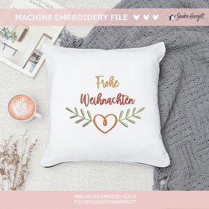 May include: White square pillow with the embroidered text "Frohe Weihnachten" in gold and red, with a heart and leaf design. The pillow is on a neutral surface with a cup of coffee and a book nearby.