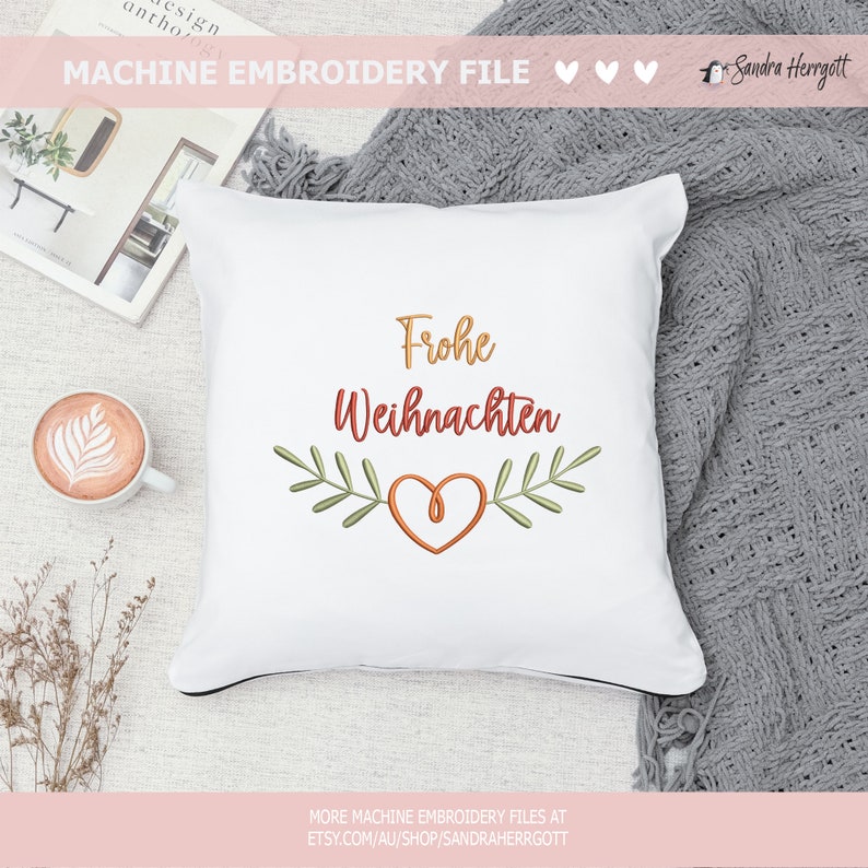 May include: White square pillow with the embroidered text "Frohe Weihnachten" in gold and red, with a heart and leaf design. The pillow is on a neutral surface with a cup of coffee and a book nearby.