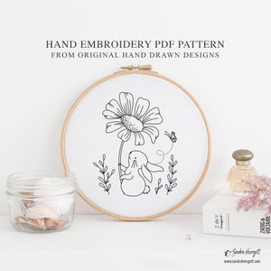 Op de afbeelding: Een handborduurpatroon met een zwart-witte lijntekening van een konijn dat een bloem vasthoudt, met een vlinder. Het ontwerp is geplaatst in een houten borduurring. De tekst op de afbeelding luidt: HAND EMBROIDERY PDF PATTERN FROM ORIGINAL HAND DRAWN DESIGNS.