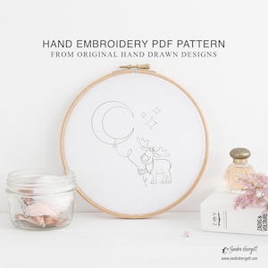 May include: A wooden embroidery hoop containing white fabric with a black line drawing of a moose holding a crescent moon balloon and stars. The text HAND EMBROIDERY PDF PATTERN FROM ORIGINAL HAND DRAWN DESIGNS is printed above the hoop.