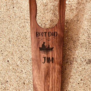 May include: A wooden boot jack with a dark brown finish. The text "BEST DAD" is above an illustration of a person and child fishing from a boat. The name "JIM" is below the illustration.