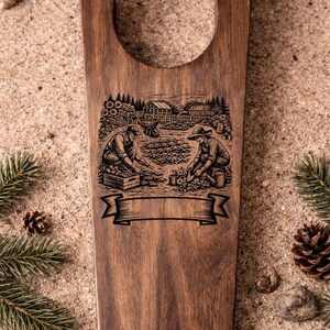 May include: A wooden boot jack with a carved illustration of two people gardening. The dark brown wood has a smooth finish. A banner is carved below the illustration. The boot jack is surrounded by pine needles, pine cones, and sand.