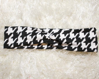 May include: Black and white houndstooth patterned headband. The headband is twisted and has a knot in the centre.