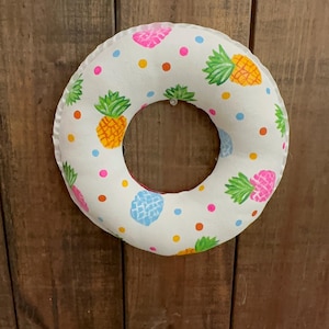 May include: A round, white fabric wreath with a repeating pattern of colorful pineapples, pink and blue shapes, and dots. The wreath is approximately 12 inches in diameter and has a circular opening in the center. The background is a wooden surface.