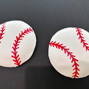 May include: Two white baseball-shaped patches with red stitching. The patches are on a black background.